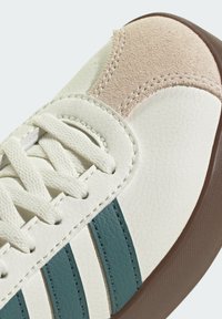 Biele kožené tenisky s jemne hnedými semišovými akcentmi, so tyrkysovými pruhmi, textúrovaným povrchom a plochou gumovou podrážkou. Šnúrky sú biele.