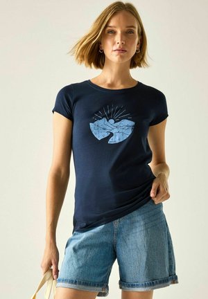 Kobieta nosząca granatowy t-shirt z niebieskim motywem góry i słońca oraz podwinięte dżinsowe szorty, trzymająca beżową torbę jedną ręką.