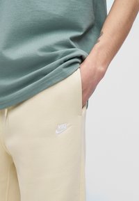 Asmuo, dėvintis smėlio spalvos Nike laisvalaikio kelnes, ranka kišenėje ir žalsvai mėlyną marškinius, rodo mažą dilbio tatuiruotę.