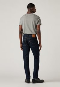 Joven de pie con una camiseta gris, jeans azul oscuro con bolsillos traseros y un parche de cuero, y zapatos negros, contra un fondo liso.