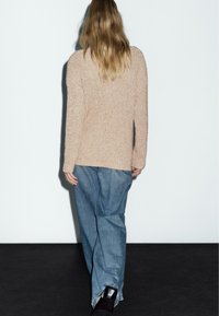 Beige stickad tröja med en texturerad yta, bärs med lösa blå denimbyxor. Baksidan har en enkel, ren design.
