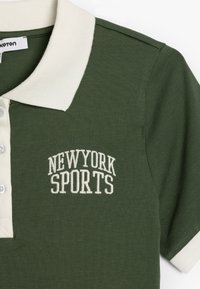 Grøn polo skjorte med hvid ribbet krave, tre knapper og broderet "NEWYORK SPORTS" tekst i hvidt. Blødt stof med en tekstureret overflade.