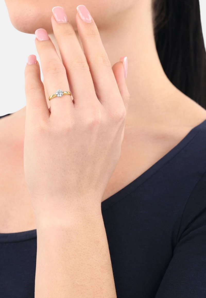 Anello d'oro con una pietra preziosa rotonda e trasparente. La mano è posizionata delicatamente, con un tono di pelle chiaro e unghie curate.