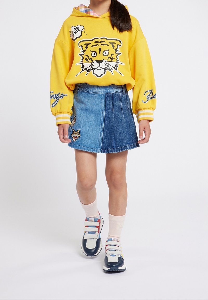 Hoodie jaune avec un graphique de tigre et des détails blancs, associé à une jupe en denim bleu en forme de A avec des patchs et un ourlet brut. Baskets blanches et marine.