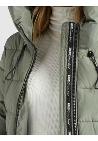 Puffy olive green jacket with a hood, featuring a black zipper and branded tape detail along the front, worn over a light ribbed turtleneck.