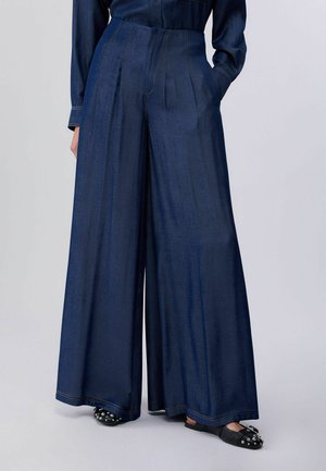 Pantaloni di denim blu scuro a gamba larga con pieghe frontali, indossati con scarpe slip-on nere con borchie, mostrati dalla vita fino ai piedi.