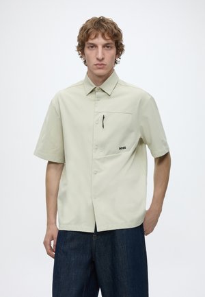 Jeune homme aux cheveux bouclés portant une chemise beige clair à manches courtes avec poche poitrine et un jean foncé sur un fond uni.