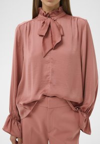 Lyserød silke bluse med en høj, flæset krave og sløjfedetalje. Lange ærmer med samlede manchetter. Glat tekstur og let løs pasform.