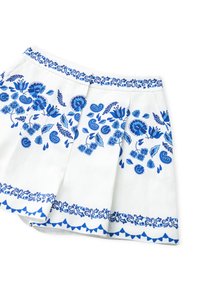 Gonna bianca con ricami floreali blu. Presenta una vita decorativa e un orlo con un motivo triangolare. Il design plissettato aggiunge texture.