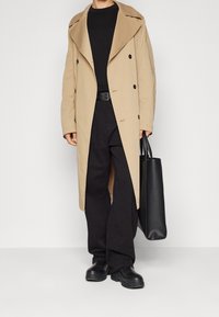 Béžový dlouhý kabát s velkým límcem, dvouřadovými knoflíky, nošený přes černý top a černé kalhoty, doplněný černou taškou přes rameno a masivními botami.