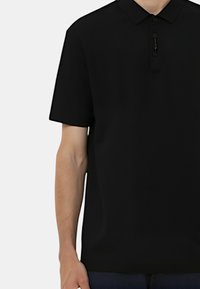 Polo shirt corto nero a maniche corte con colletto classico, placca con due bottoni e texture liscia, indossato con jeans scuri.