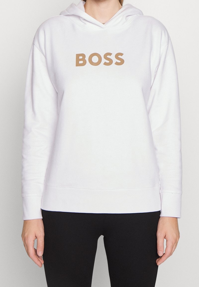 Osoba ubrana w biały kaptur z dużym napisem "BOSS" w kolorze beżowym na piersi i czarne spodnie, stojąca na prostym tle.