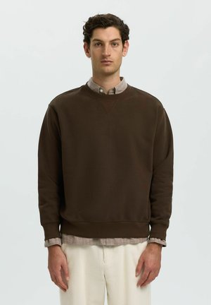 Jeune homme aux cheveux courts, bouclés et foncés, portant un sweat-shirt marron par-dessus une chemise beige et un pantalon clair, debout devant un fond uni.
