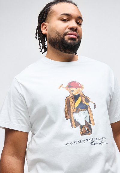 Άνδρας με πλεγμένα μαλλιά που φοράει λευκό μπλουζάκι με την απεικόνιση του Polo Bear ντυμένου με εξοπλισμό ιππασίας και κρατώντας ματσόλα πόλο.