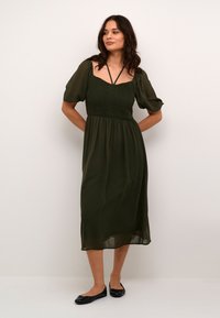 Rochie midi de culoare verde închis cu un bust elastic, mâneci bufante și decolteu pătrat. Materialul pare ușor și fluid. Pantofi negri purtați.