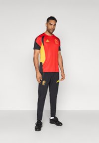 Crveno-crna atletska odjeća s žutim detaljima; majica kratkih rukava s teksturiranim dizajnom i logotipom; crne hlače u kompletu s logotipom.