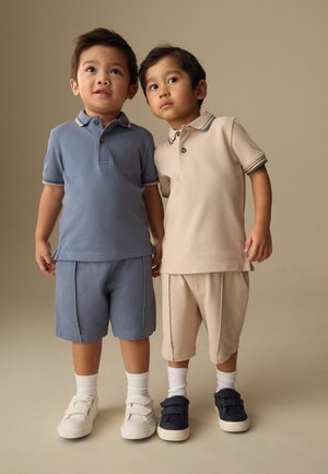 Two young boys stand side by side, wearing matching polo shirts and shorts, one in blue and the other in beige, with white socks and sneakers.