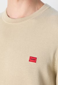 Beige Strickpullover mit einem roten rechteckigen Patch mit der Aufschrift "HUGO" auf der Brust, getragen von einer Person mit sichtbarem Hals und Schulter.