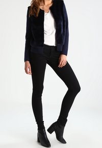 Granatowa kurtka bomber z futerkiem typu faux fur z ściągaczami na mankietach, nałożona na białą bluzkę, zestawiona z czarnymi obcisłymi jeansami i czarnymi botkami na obcasie.