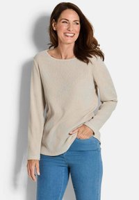 Lächelnde Frau mit langen braunen Haaren, die ein beiges, langärmliges, strukturiertes Oberteil und eine blaue Jeans trägt, steht vor einem weißen Hintergrund.
