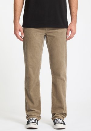 Homme portant un pantalon en velours beige, un T-shirt noir et des baskets noires avec des lacets blancs, debout devant un fond blanc.