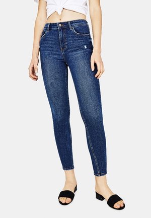 Kvinde iført mørkeblå skinny jeans, hvid knyttet top der viser maven, og sorte lavhælede åbne tå-sandaler, stående mod en hvid baggrund.