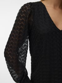 Μαύρη δαντελένια μπλούζα με διαφανείς μακρύες μανίκες με σχέδιο και V-λαιμό, πάνω σε ένα άτομο, σε απλή φόντο.