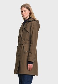 Femme portant un trench-coat marron ceinturé avec des boutons et un pull sombre en dessous, debout devant un fond clair uni.