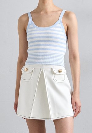 Frau trägt ein ärmelloses, hellblau-weiß gestreiftes Crop-Top kombiniert mit einem weißen Minirock mit Fronttaschen und goldenen Knöpfen.