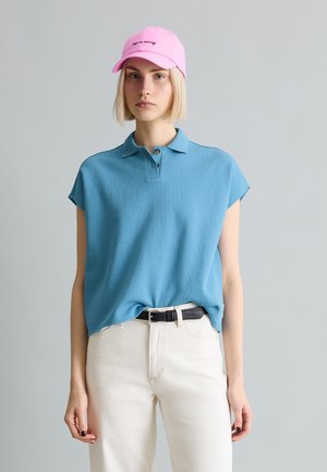 Kobieta w różowej czapce, niebieskiej koszulce z krótkim rękawem z kołnierzykiem, białych spodniach i czarnym pasku na tle jednolitego szarego tła.