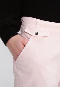 Ljusrosa byxor i texturerad tyg med en framficka och metallknappstängning, visade med en hand vilande i fickan.