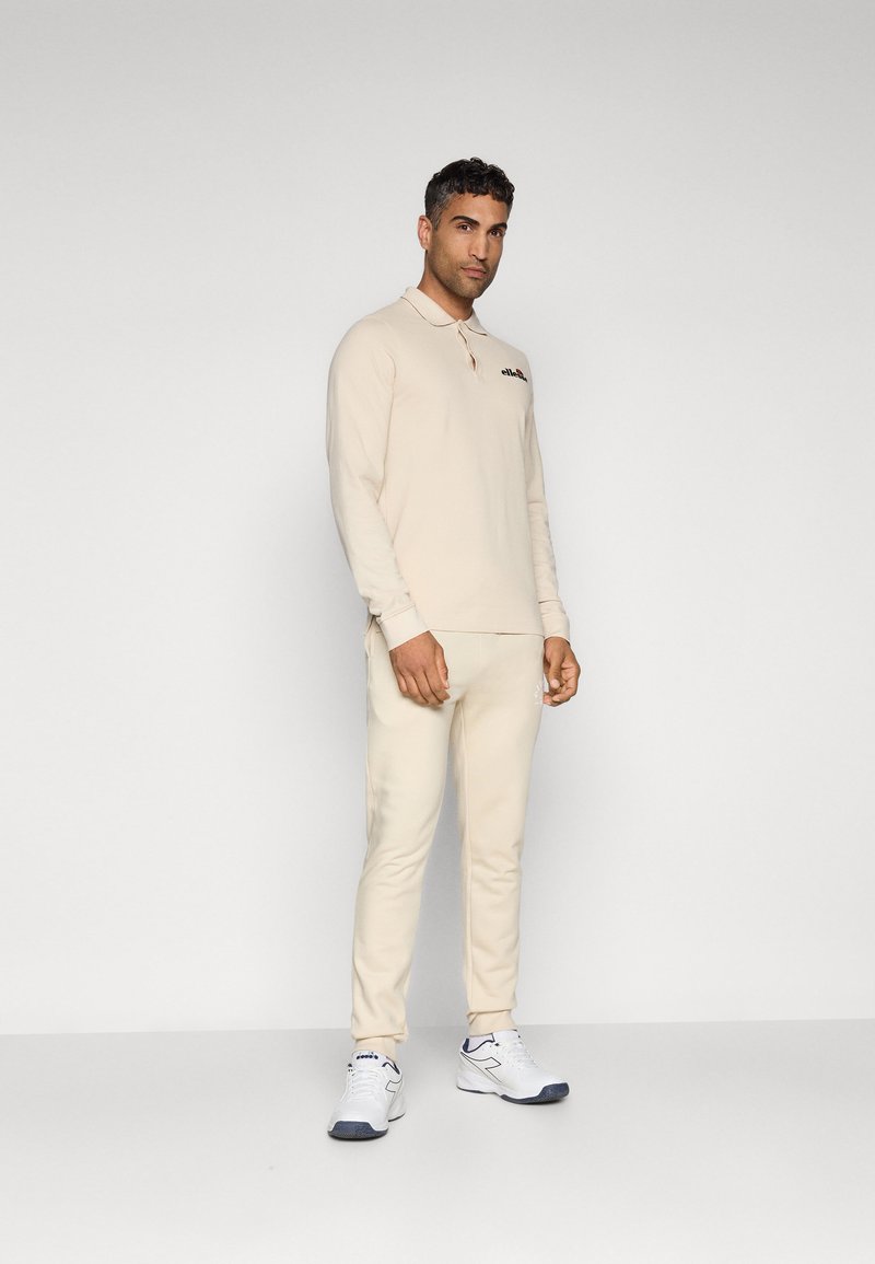 Beige Langarmpolo mit kleinem Logo, kombiniert mit passenden beigen Hosen und weißen Sneakers mit blauen Akzenten. Glatte Textur im gesamten Design.