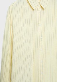 Rumena srajca z dolgimi rokavi, gumbi na sprednji strani, z ozkimi vertikalnimi rjavimi črtami in klasičnim ovratnikom, prikazana od spredaj.