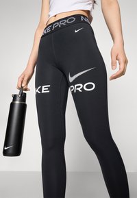 Schwarze Nike Pro Leggings mit weißen Logo-Details, einschließlich einem hohen Bund und glattem, dehnbarem Material. Eine schwarze Wasserflasche wird in der Hand gehalten.
