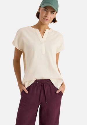 Młoda osoba nosząca kremowy teksturowany krótki top, bordowe spodnie ze sznurkiem i zieloną czapkę z daszkiem, stojąca z rękami w kieszeniach.