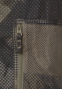Närbild av ett brunt och svart prickigt tyg med en nätficka och en synlig YKK-dragkedja på vänster sida.