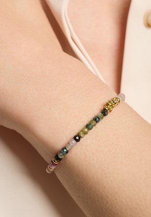 Bracelet en perles avec des pierres précieuses vertes, noires, bleues et roses, entrecoupées d'accents dorés, porté sur un poignet à la peau claire.