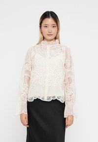 Blusa de encaje en suave rosa pálido con patrones florales, cuello ruffled y mangas acampanadas, combinada con una falda negra lisa.