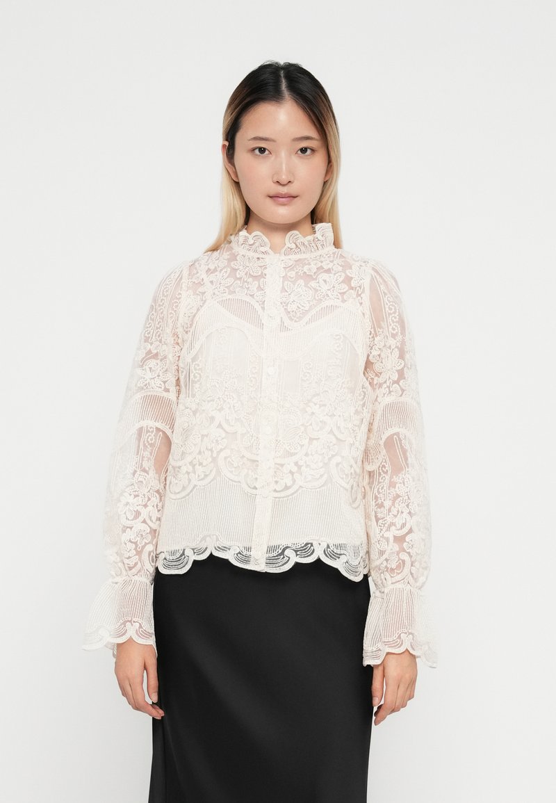Blusa de encaje en suave rosa pálido con patrones florales, cuello ruffled y mangas acampanadas, combinada con una falda negra lisa.