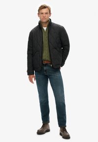 Chaqueta acolchada negra, suéter de punto trenzado verde, jeans azules y botas marrones con cordones. Presenta un cuello alto y texturas contrastantes.