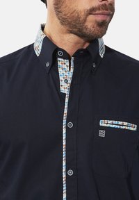 Marineblaues Hemd mit Knöpfen, das einen gemusterten Kragen und eine Taschenakzent hat, in einem Hahnentrittdesign in Blau-, Braun- und Weißtönen.