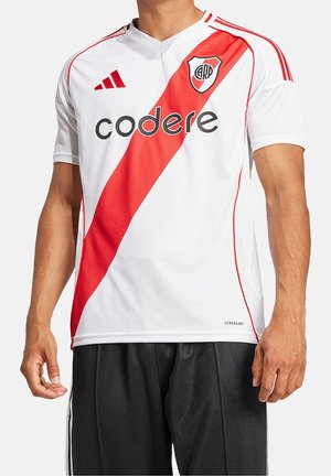 Mężczyzna w białej koszulce piłkarskiej z czerwonym ukośnym paskiem, logo River Plate, logo Adidas i nazwą sponsora „codere”, w połączeniu z czarnymi szortami.