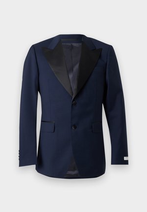 Man staat met handen in de zakken, gekleed in een donkerblauw pak met zwarte revers, een wit overhemd en bruine nette schoenen, tegen een effen lichte achtergrond.