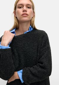 Ciemny, teksturowany sweter z szerokimi rękawami, nałożony na jasnoniebieską koszulę z kołnierzykiem. Materiał wydaje się puszysty, co nadaje mu przytulny wygląd.