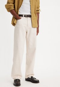 Licht beige wijde broek met een lichte textuur, gecombineerd met een bruine gevlochten leren riem en zwarte leren schoenen. Olijfgroene button-up shirt.