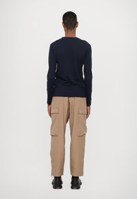 Jūrų laivyno ilgomis rankovėmis marškiniai su prigludusiu dizainu, derinami su smėlio spalvos krovinių kelnėmis su šoninėmis kišenėmis ir juodais tekstūruotais batais.