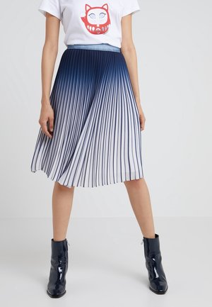 Femme portant un t-shirt blanc avec un motif de visage de chat rouge, une jupe midi plissée bleu et blanc dégradé, et des bottines navy brillantes.