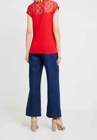 Donna che indossa una maglietta rossa a maniche corte con dettagli in pizzo sulla schiena, jeans blu a gamba larga e sandali con tacco beige, in piedi di spalle.