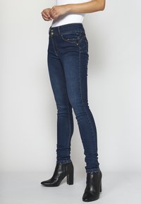 Jeans ajustados de mezclilla en azul oscuro, con una cintura alta, bolsillos frontales y costuras en contraste. Llevados con botas negras hasta el tobillo.