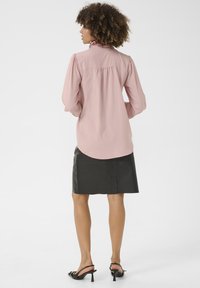 Camicia rosa chiaro con colletto arricciato, maniche lunghe a sbuffo e vestibilità ampia. Abbinata a una gonna in pelle nera e tacchi slingback neri.